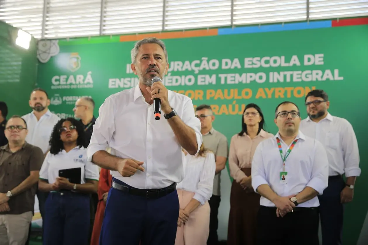 O governador Elmano de Freitas fala ao microfone à frente de um painel verde que anuncia a inauguração da Escola de Ensino Médio em Tempo Integral Professor Paulo Ayrton. Ele está cercado por secretários e assessores durante a cerimônia.