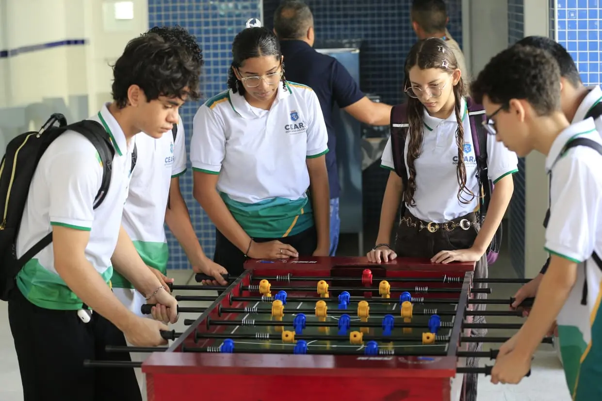 Quatro estudantes com o uniforme da rede estadual do Ceará se divertem jogando totó em um pátio escolar. Eles estão concentrados na partida em um ambiente de convivência com paredes de azulejos azuis.