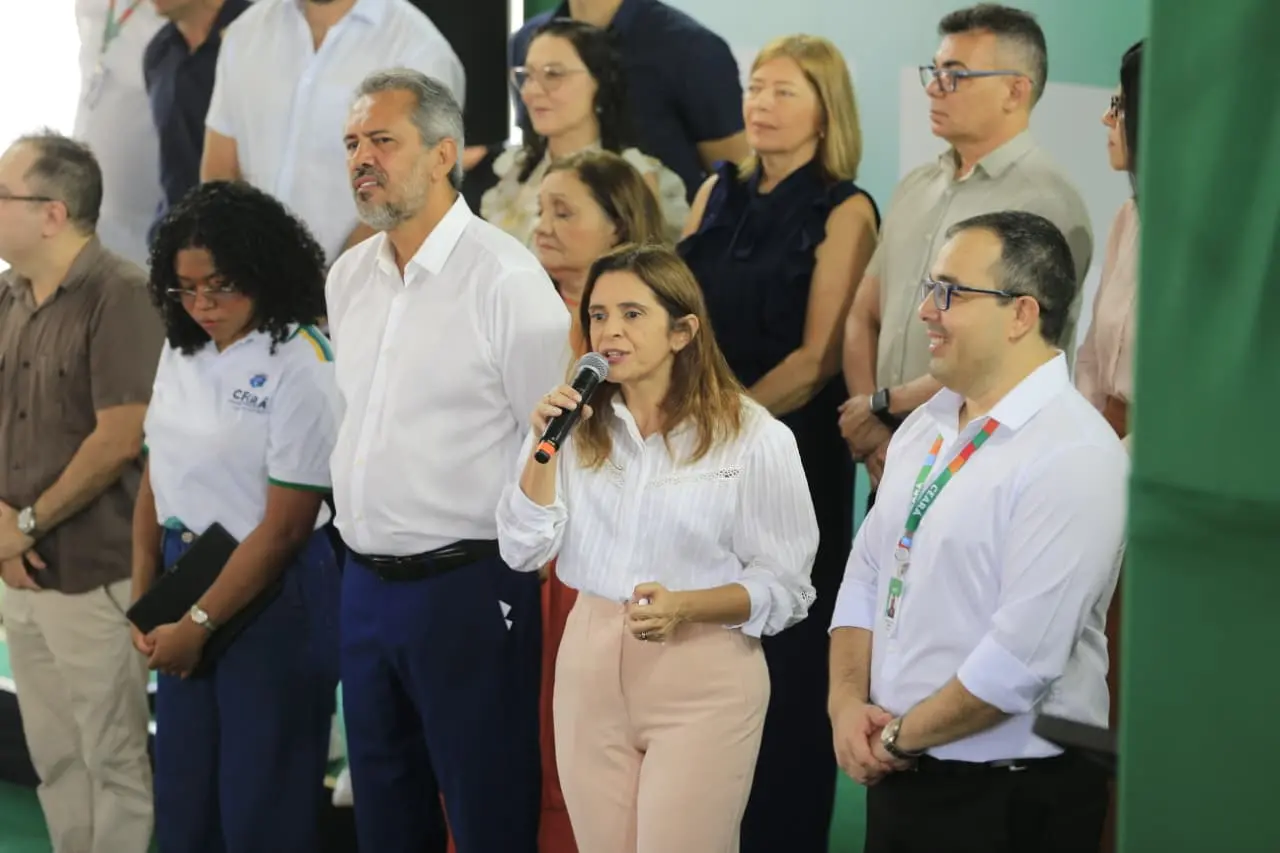 A Secretária da Educação do Ceará, Eliana Estrela, discursa ao microfone durante um evento oficial. Ela veste uma blusa branca e calça clara, acompanhada pelo governador Elmano de Freitas e outras autoridades ao fundo.