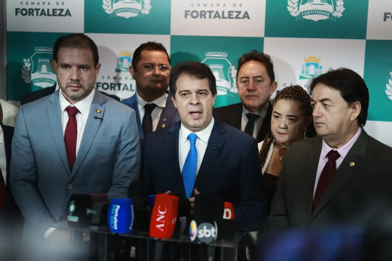 Grupo de autoridades em coletiva de imprensa na Câmara Municipal de Fortaleza, posicionadas atrás de um púlpito com vários microfones de emissoras de TV. Ao fundo, painel institucional com o brasão e o nome da Câmara de Fortaleza.