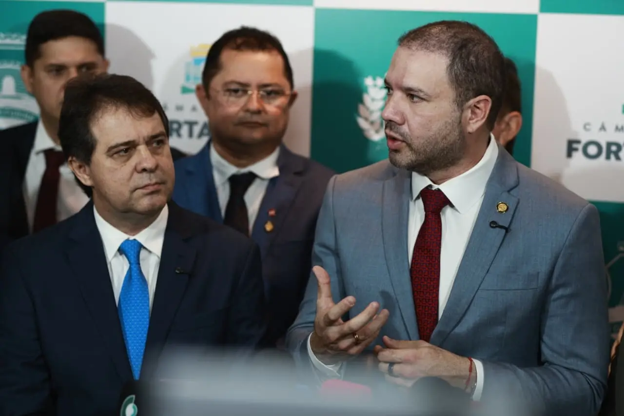 Dois homens de terno em primeiro plano durante coletiva de imprensa na Câmara Municipal de Fortaleza; um deles fala gesticulando enquanto o outro o observa. Ao fundo, outras autoridades e painel institucional da Câmara.