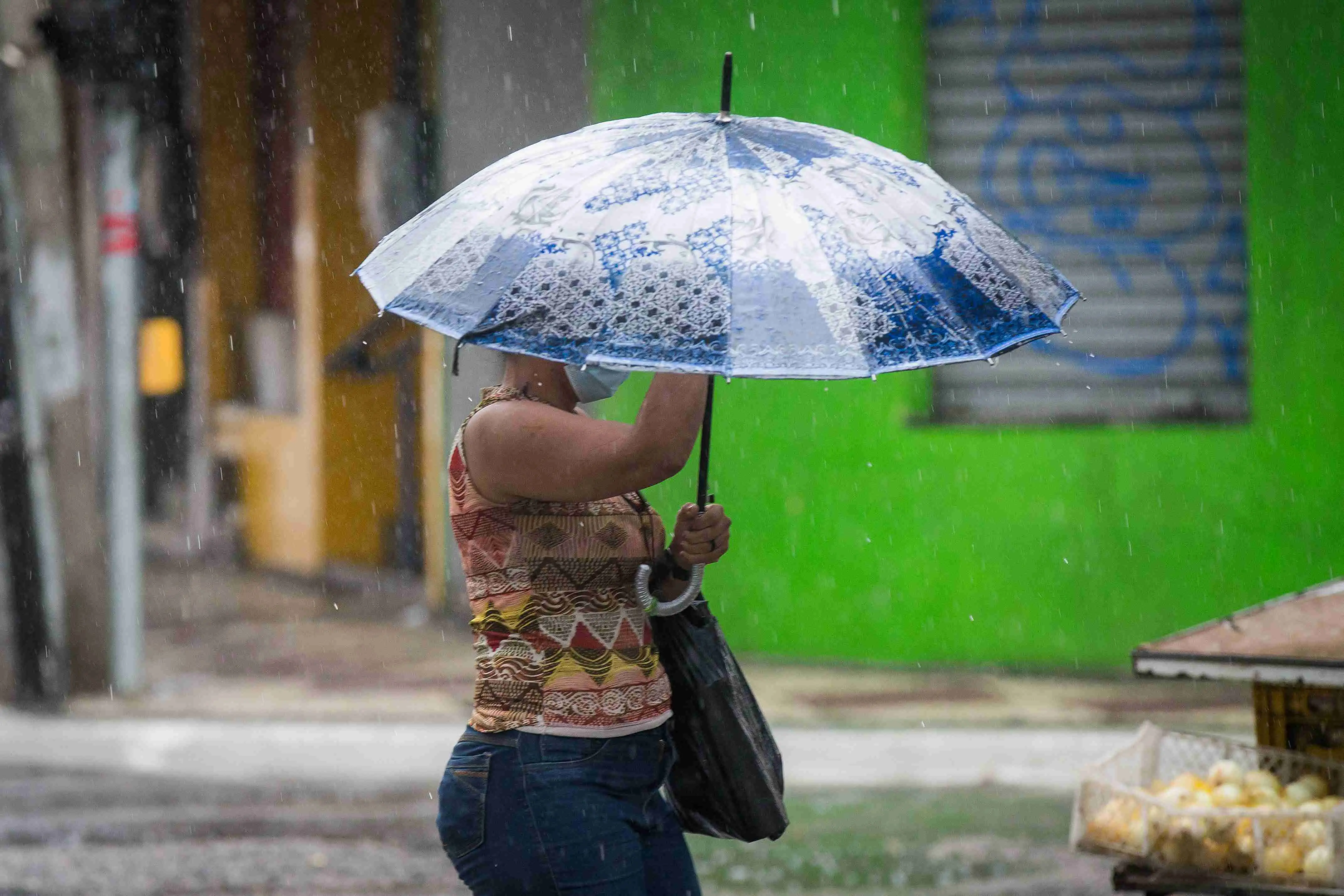 Mulher protegida por guarda-chuva durante um dia chuvoso, com edifícios coloridos ao fundo.