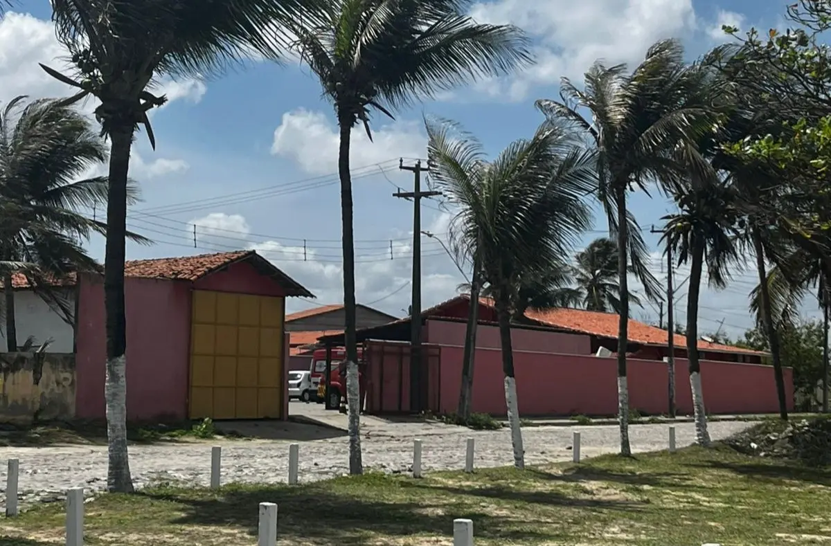 Rua costeira com palmeiras altas, casas de telhado vermelho e um céu azul parcialmente nublado. A casa é a sede do Quartel dos Bombeiros em Icaraí, no Ceará.