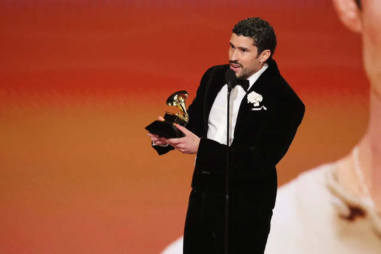 Pessoa usando terno preto com camisa branca, segurando um troféu do Grammy enquanto fala ao microfone, sobre um palco iluminado em tons quentes.