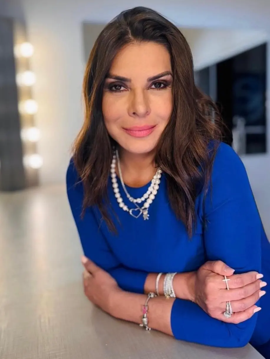 Pessoa com cabelos longos e castanhos, vestindo um vestido azul vibrante, apoiada sobre uma superfície clara. Ela usa vários acessórios prateados, incluindo colares, pulseiras e anéis. O ambiente ao fundo é interno, com iluminação suave e luzes de camarim desfocadas.