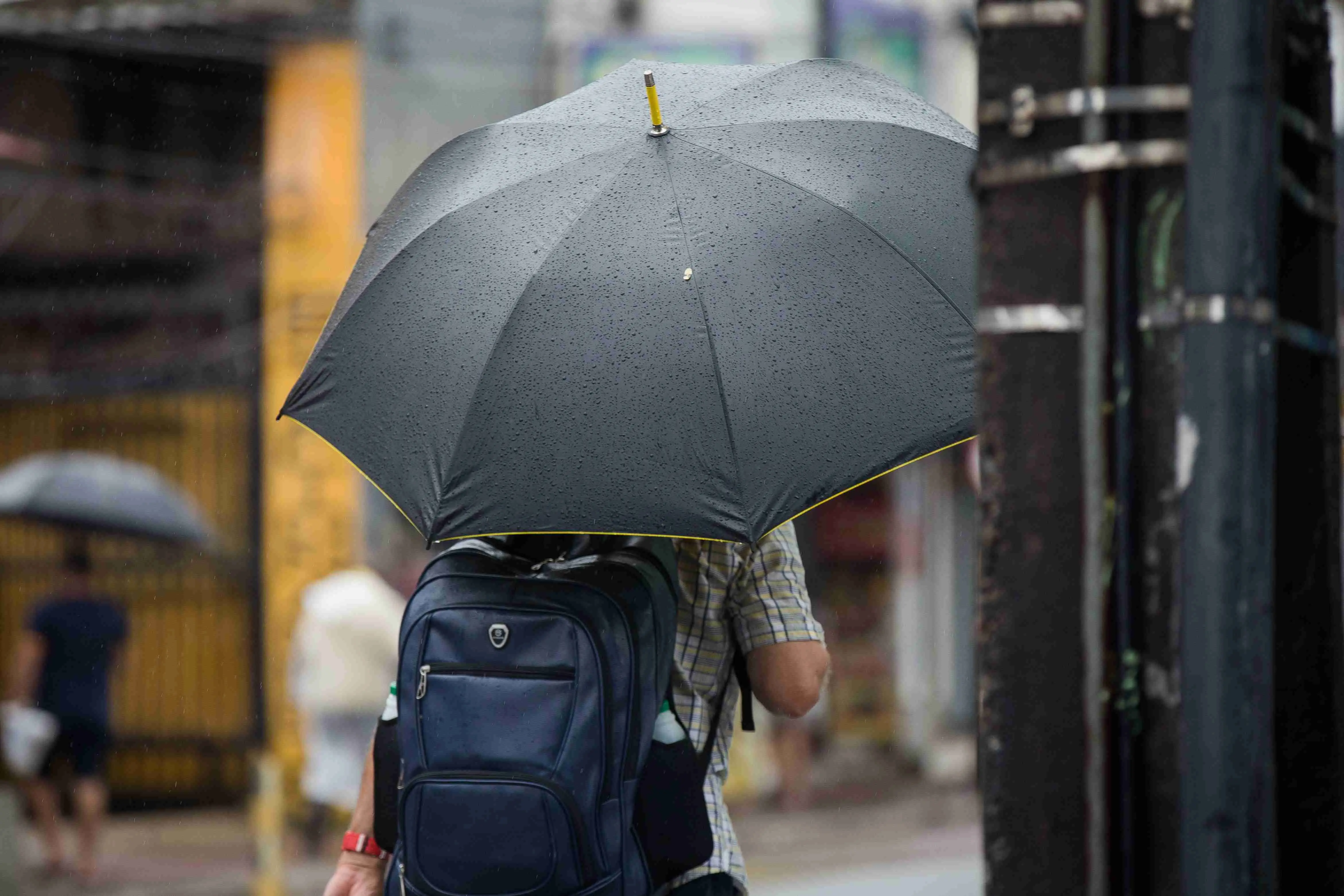 Homem caminhando em dia chuvoso, vista por trás, segurando um guarda-chuva preto coberto de gotas de chuva e usando uma mochila azul.