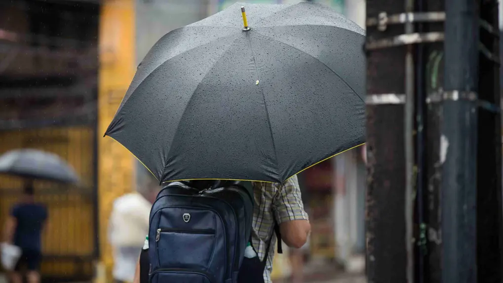 Homem caminhando em dia chuvoso, vista por trás, segurando um guarda-chuva preto coberto de gotas de chuva e usando uma mochila azul.