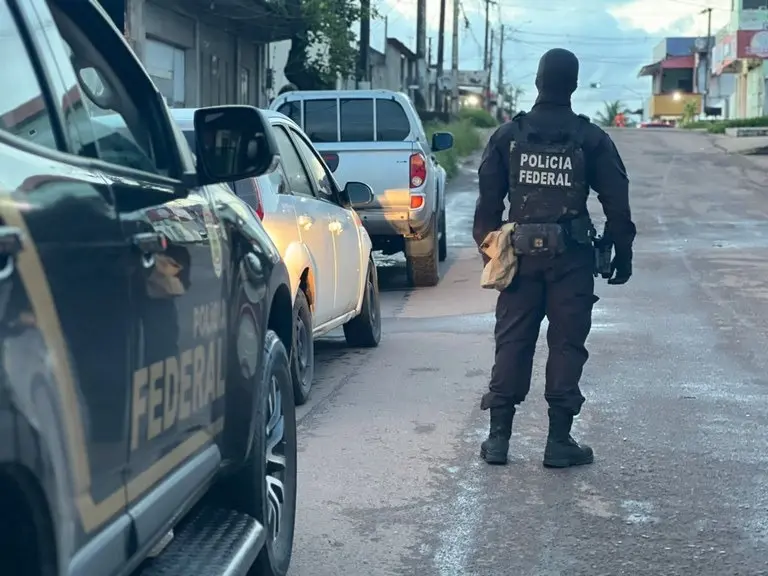 A foto mostra um policial federal de costas, fardado e com balaclava, em uma rua onde há uma viatura da Polícia Federal e outros dois carros estacionados.