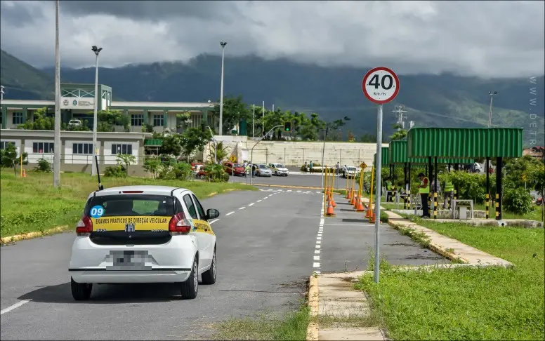 Um carro branco de autoescola com a inscrição 'EXAMES PRÁTICOS DE DIREÇÃO VEICULAR' trafegando em uma pista de testes do DETRAN, com cones de sinalização, uma placa de limite de velocidade de 40 km/h e montanhas nebulosas ao fundo.