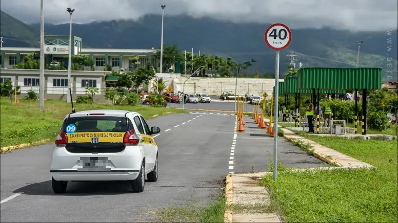 Um carro branco de autoescola com a inscrição 'EXAMES PRÁTICOS DE DIREÇÃO VEICULAR' trafegando em uma pista de testes do DETRAN, com cones de sinalização, uma placa de limite de velocidade de 40 km/h e montanhas nebulosas ao fundo.