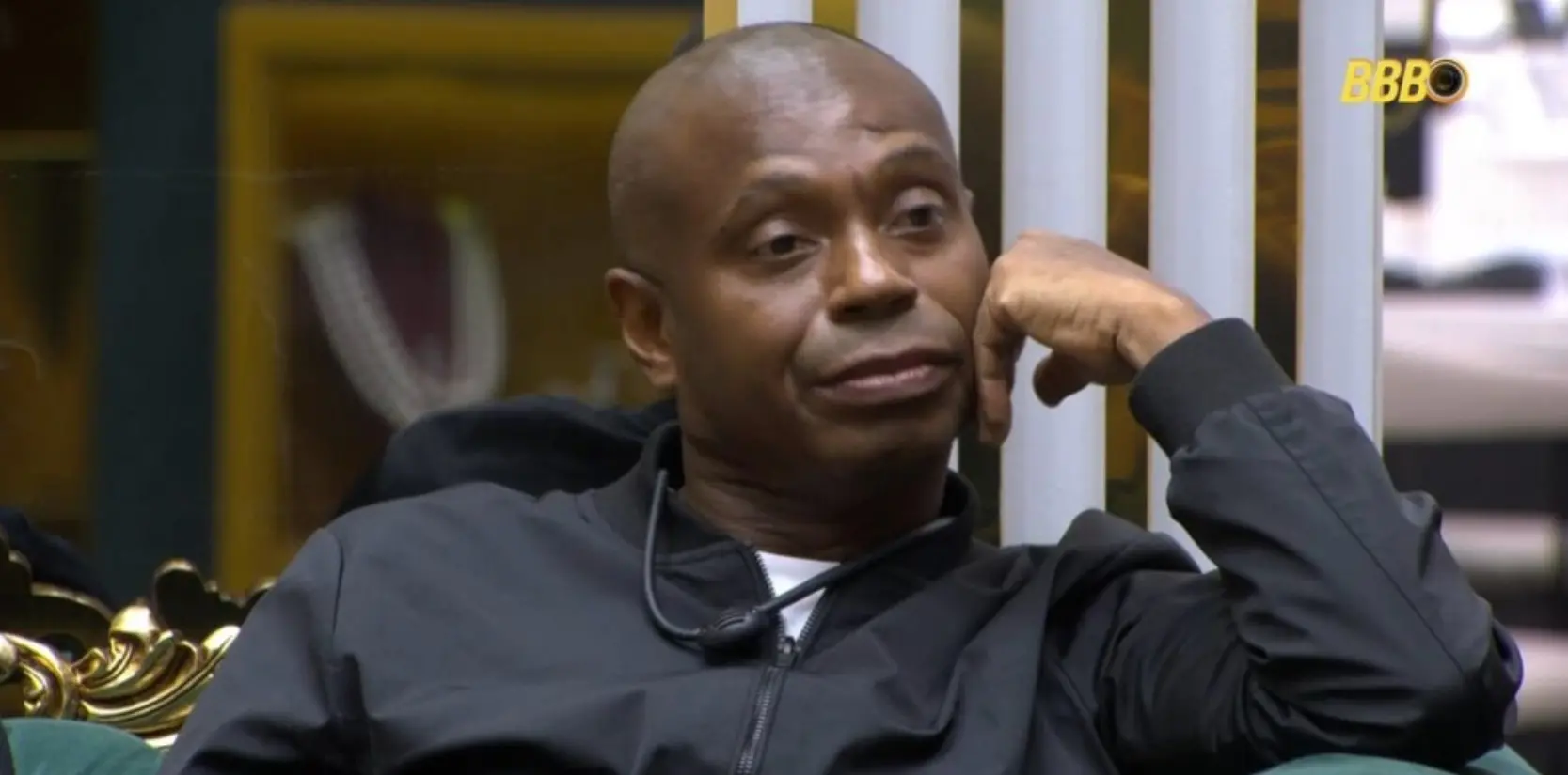 Edílson Capetinha no BBB 26 com expressão pensativa, sentado em um ambiente interno, vestindo uma jaqueta preta, com fundo que inclui uma estante dourada e uma parede branca.