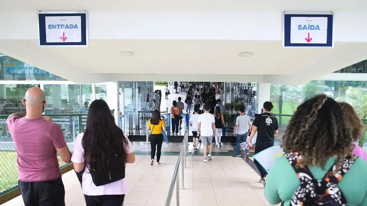 Imagem mostra grupo de pessoas caminhando em um corredor moderno com placas de ENTRADA e SAÍDA visíveis em uma universidade.