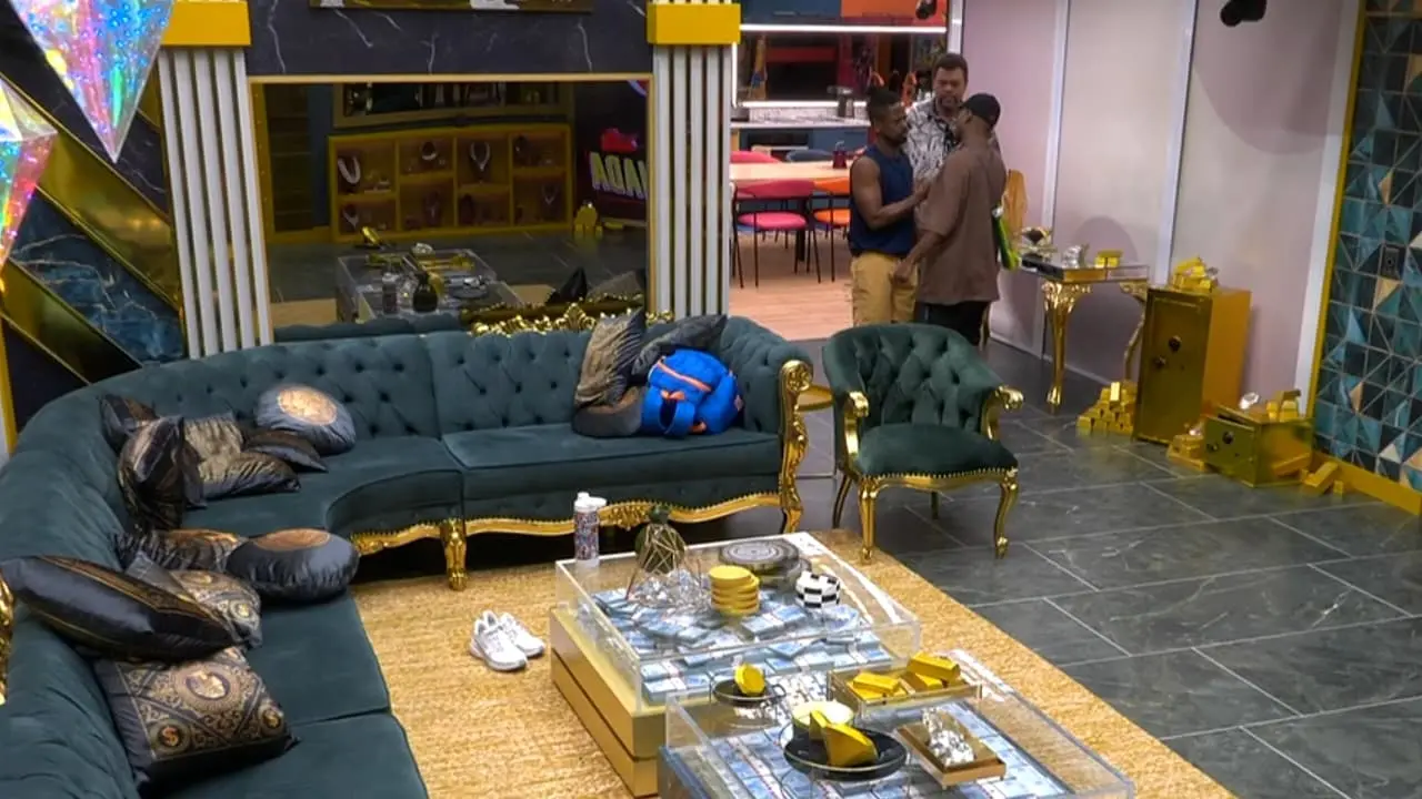 Sala ampla e decorada com sofás verde-esmeralda de estilo clássico com detalhes dourados, várias almofadas estampadas e uma mesa de centro transparente repleta de objetos dourados e itens organizados. Há um tapete grande em tom de palha cobrindo parte do chão, que combina com o piso de mármore escuro ao redor. Ao fundo, três pessoas estão em pé conversando perto da entrada para outra área da casa.