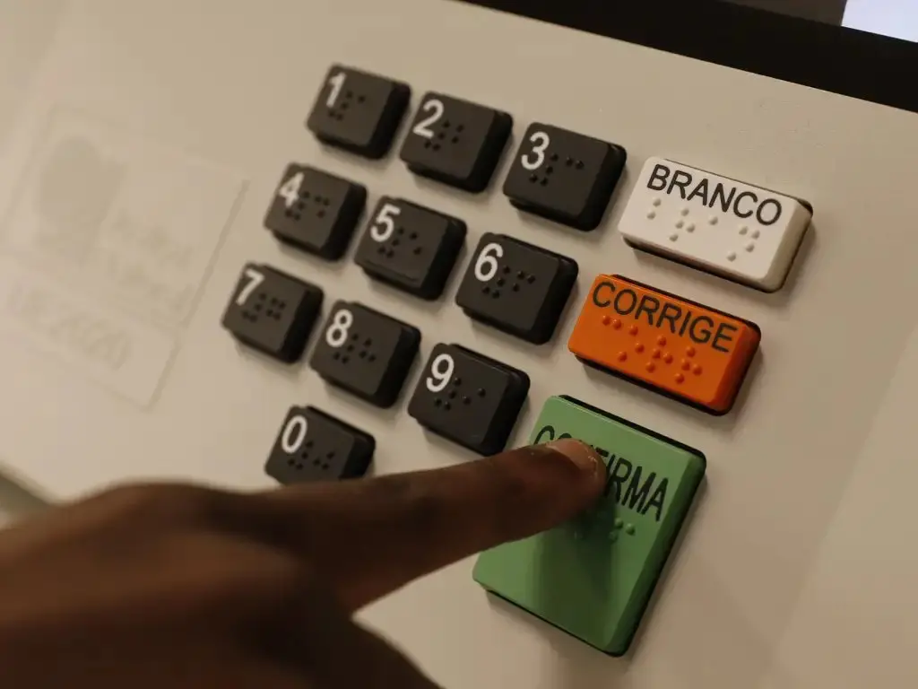 Máquina branca com números de 0 a 9 e botões de Branco, Corrige e Confirma, com dedo indicador negro clicando na opção Confirma.