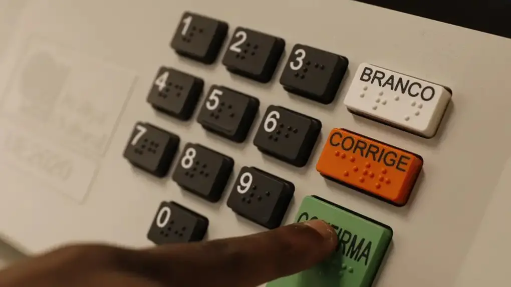 Máquina branca com números de 0 a 9 e botões de Branco, Corrige e Confirma, com dedo indicador negro clicando na opção Confirma.