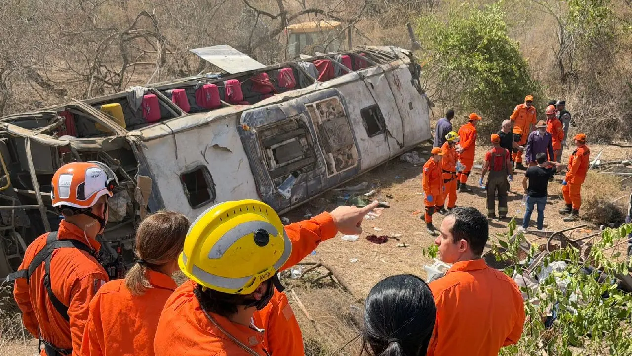 Fotografia em plano médio mostrando o local de um grave acidente de ônibus em uma área de vegetação seca. Um ônibus branco e cinza está tombado de lado, com o teto parcialmente destruído, revelando poltronas vermelhas no interior. Vários socorristas do Corpo de Bombeiros, vestindo uniformes laranja vibrante e capacetes de proteção, estão espalhados pela cena. Em primeiro plano, as costas de quatro socorristas são visíveis, com um deles apontando para o veículo. Ao fundo, outros bombeiros e civis se agrupam próximos à traseira do ônibus em uma área de terra batida e galhos secos. O dia está ensolarado.