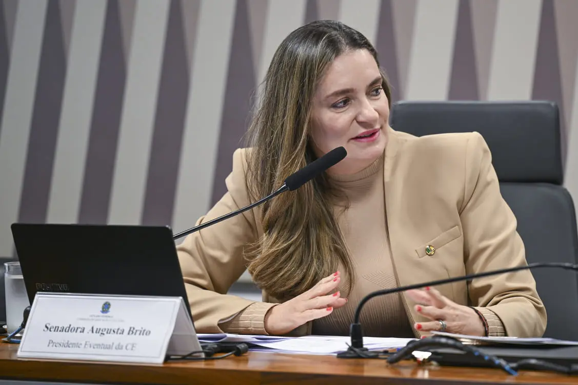 Augusta Brito, mulher branca de cabelos loiros e lisos, usando terno bege e falando em microfone enquanto está sentada na cadeira de uma bancada.
