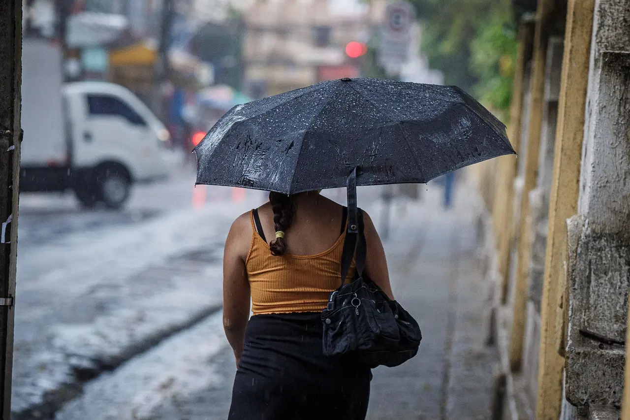 Mulher de costas com blusa amarela e trança, segurando um guarda-chuva molhado enquanto caminha em um dia chuvoso na cidade.