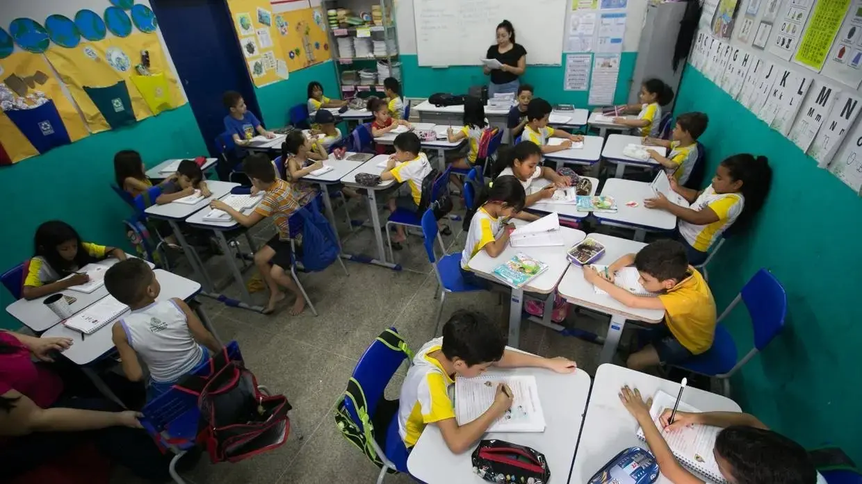 Sala de aula, com crianças sentadas em cadeiras, com mesas à frente. Professor aparece ao fundo da sala, escrevendo na lousa.