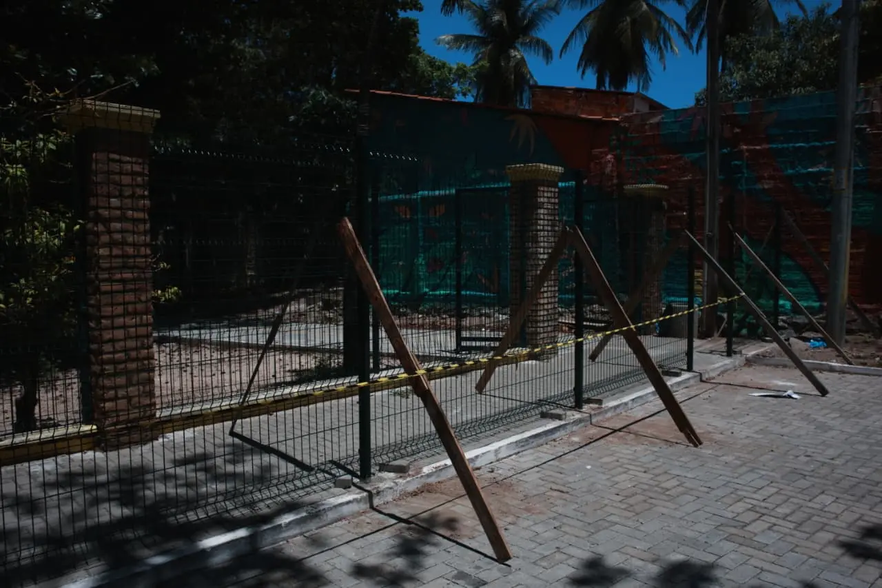 Grade metálica recém-instalada com pilares de tijolos e suportes de madeira temporários, em uma área externa pavimentada com fita de advertência amarela. Ao fundo, um muro decorado com murais coloridos e palmeiras sob céu azul.