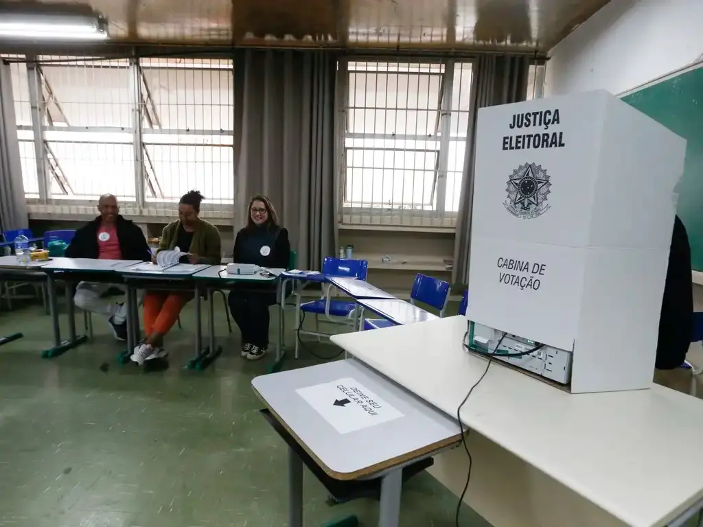 Cabine de votação em sala de aula com três fiscais sentados ao fundo.