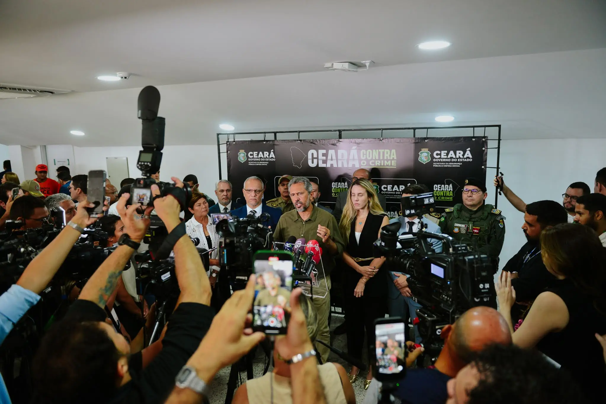A foto mostra autoridades do Governo do Ceará participando de uma coletiva de imprensa, com a presença de jornalistas e fotógrafos.
