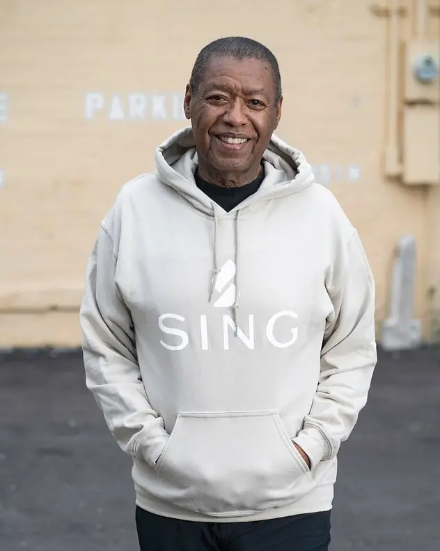 Homem sorridente de pele escura vestindo um moletom creme com capuz e o logo 'SING', com as mãos nos bolsos e uma parede bege com a palavra 'PARKING' ao fundo.