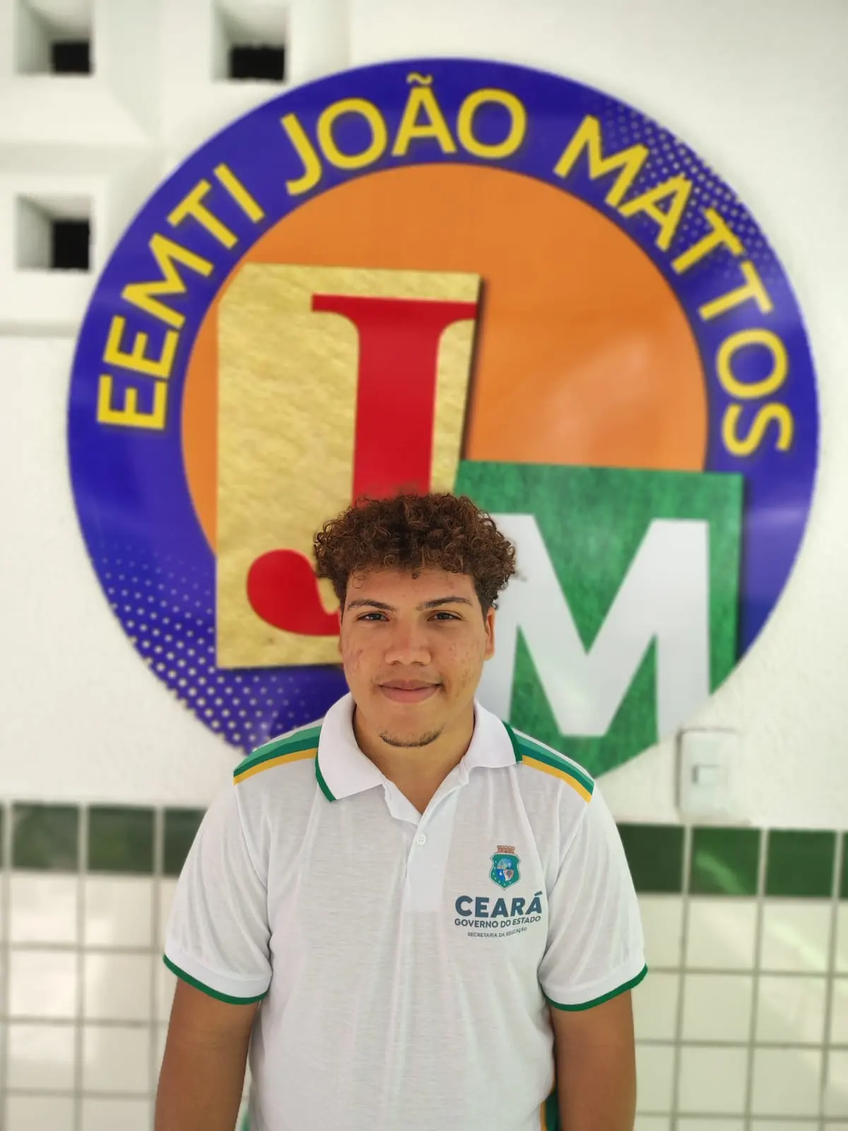 Rapaz de cabelo cacheado posando contra um emblema azul e laranja da escola EEMTI João Mattos, vestindo uma camisa polo branca, fardamento de escola estadual.