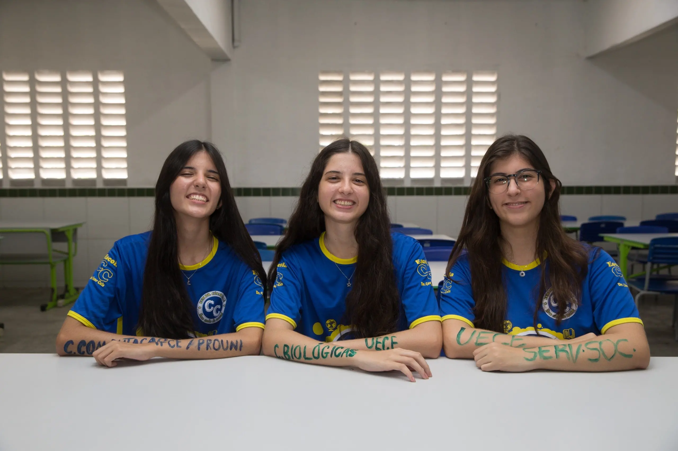Foto das trigêmeas Yasmin, Beatriz e Letícia Oliveira, jovens brancas de longos cabelos pretos, em uma sala de aula da Escola Dr. César Cals. Sentadas lado a lado, elas posam com os braços apoiados sobre uma mesa escolar, exibindo  os nomes de seus respectivos cursos pintados em tinta colorida sobre a pele.