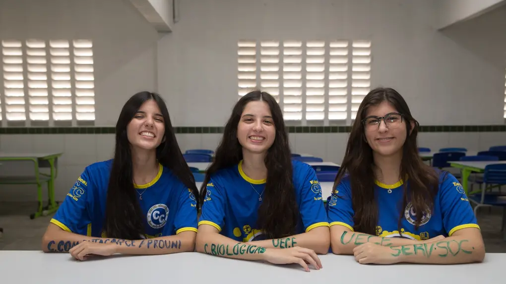Foto das trigêmeas Yasmin, Beatriz e Letícia Oliveira, jovens brancas de longos cabelos pretos, em uma sala de aula da Escola Dr. César Cals. Sentadas lado a lado, elas posam com os braços apoiados sobre uma mesa escolar, exibindo os nomes de seus respectivos cursos pintados em tinta colorida sobre a pele.