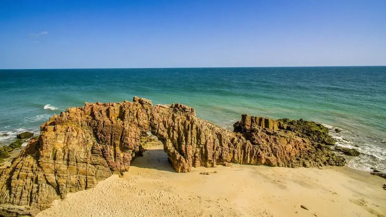 Arco natural de rocha em praia de areia dourada com mar azul-turquesa e céu claro. A visita é da Pedra Furada, em Jericoacoara, Ceará.