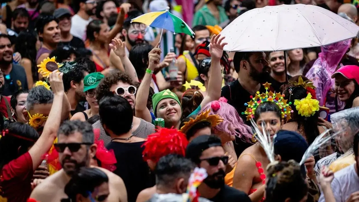 Uma fotografia colorida e vibrante captura uma multidão densa celebrando o Carnaval em uma rua de Fortaleza. No centro, pessoas usam fantasias criativas e acessórios festivos, como tiaras de girassol, bonés coloridos e óculos escuros. Destacam-se um pequeno guarda-chuva de frevo colorido aberto e um guarda-chuva branco maior à direita. O clima é de alegria e proximidade, com foliões sorrindo sob o céu nublado. A profundidade de campo é rasa, mantendo o foco nas expressões das pessoas no plano médio enquanto o fundo se mistura em um mar de cores e movimento.