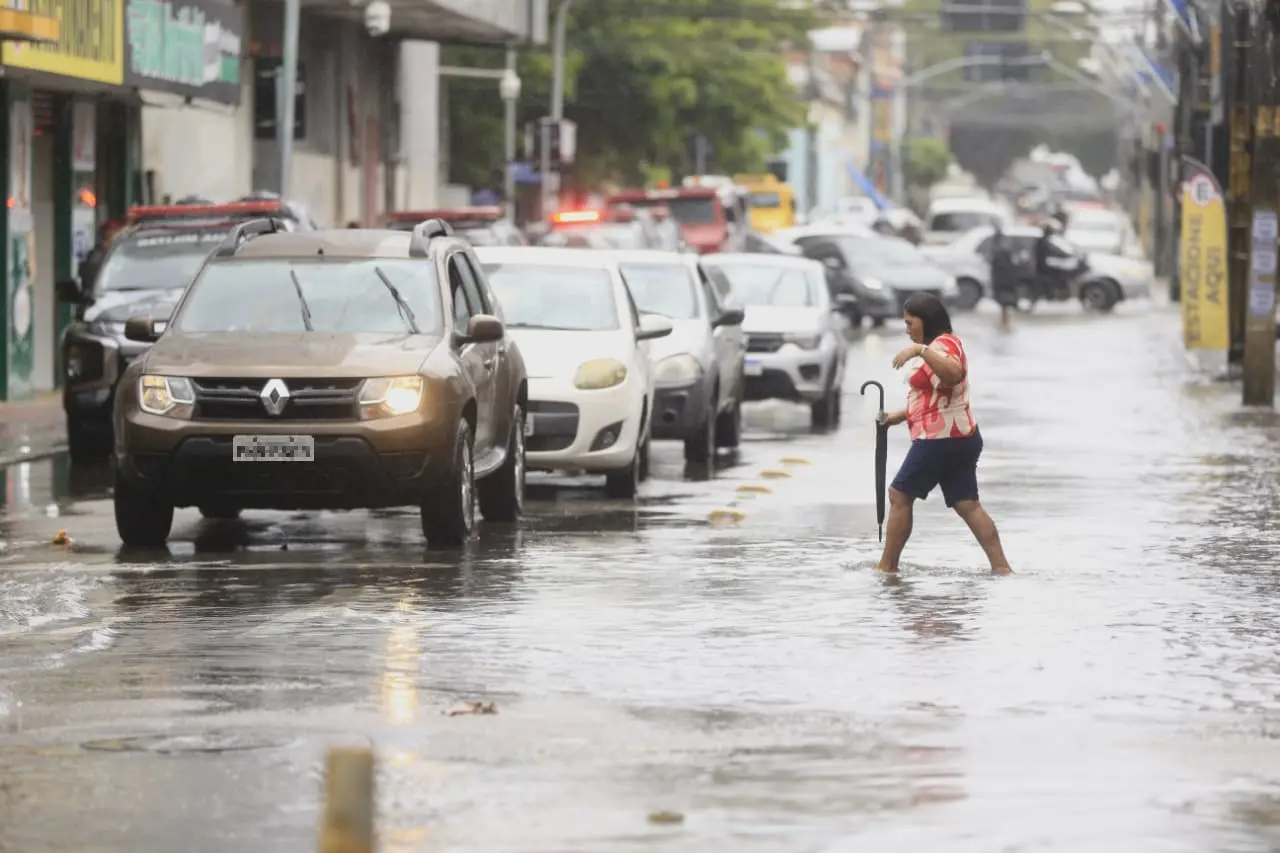 Mulher atravessando uma rua alagada e descalça, segurando um guarda-chuva, com carros parados no trânsito à sua esquerda sob chuva forte.