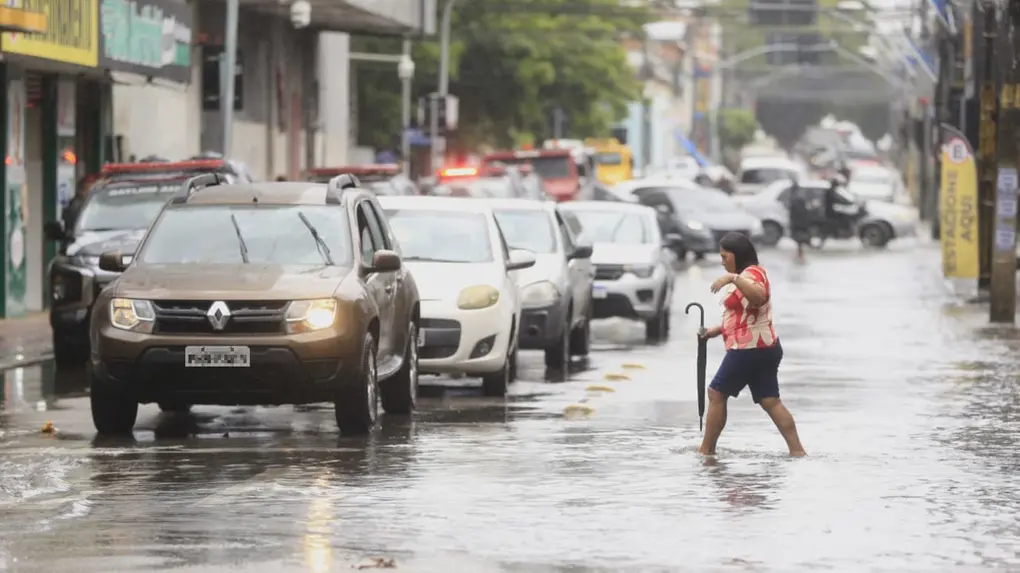 Mulher atravessando uma rua alagada e descalça, segurando um guarda-chuva, com carros parados no trânsito à sua esquerda sob chuva forte.