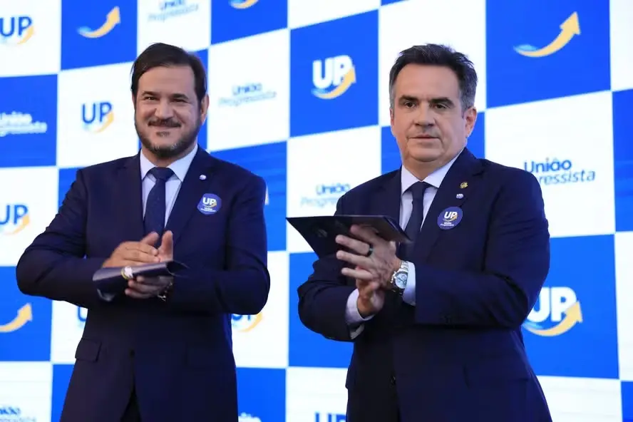 Antonio Rueda e Ciro Nogueira de ternos escuro aplaudem em um palco, diante de um painel azul e branco com o logotipo ‘UP’ e a inscrição ‘União Progressista’. Ambos usam broches no paletó e seguram tablets nas mãos.