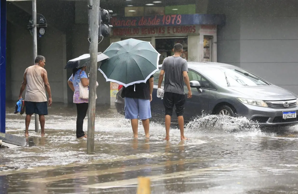 Imagem mostra pessoas em pé em uma rua alagada enquanto um carro passa e joga água nelas.