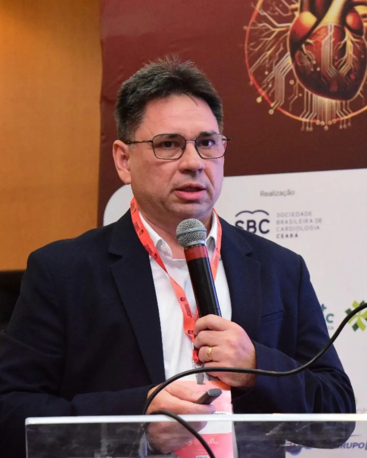 Homem de terno e óculos fala ao microfone durante palestra, em frente a painel com marcas de evento da área médica.
