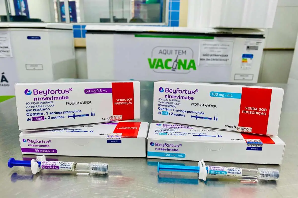 Caixas e seringas do medicamento Beyfortus (nirsevimabe), utilizadas na imunização contra o vírus sincicial respiratório (VSR), expostas em balcão de unidade de saúde com banner ‘Aqui tem vacina’ ao fundo.