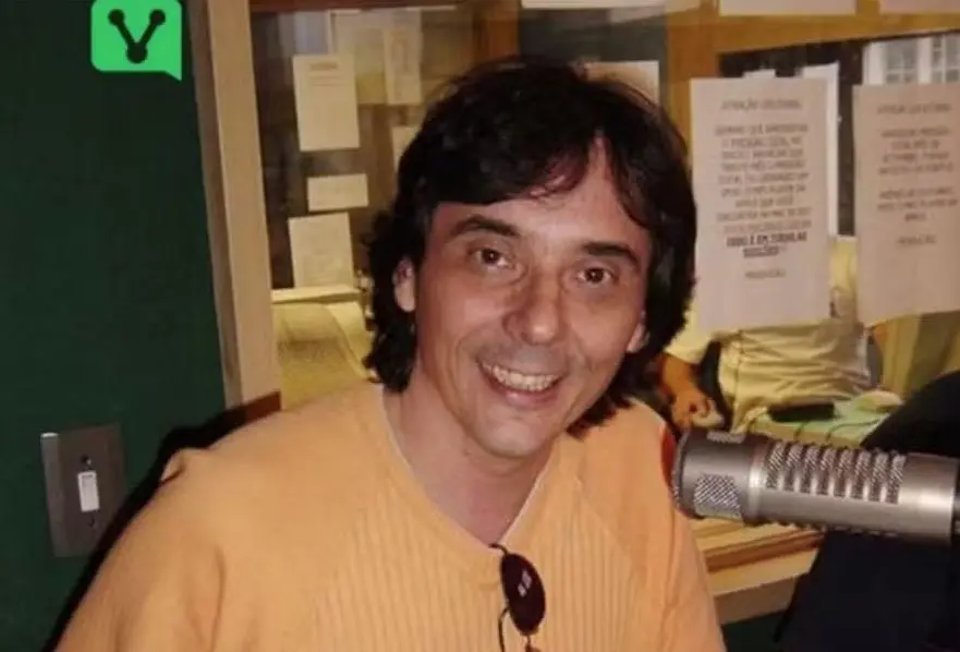 Pessoa sentada em um estúdio de rádio, à frente de um microfone profissional. A sala tem janelas de vidro ao fundo, revelando papéis colados e pessoas trabalhando em outra área do estúdio. A pessoa veste um suéter bege e tem cabelo escuro e ondulado.