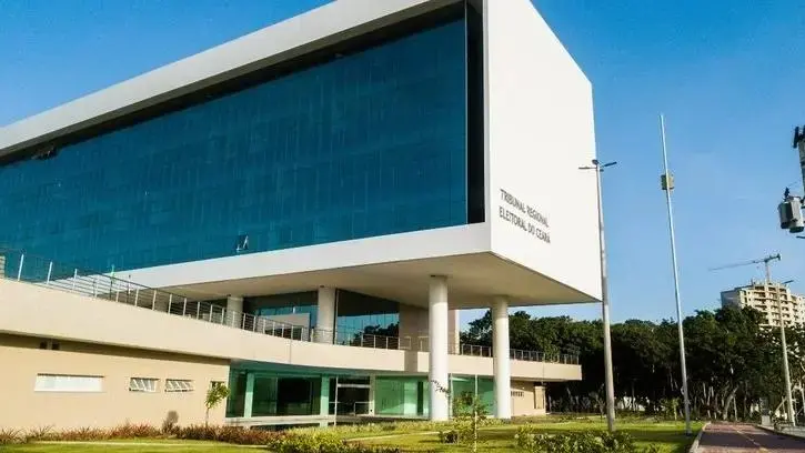 Fachada do prédio do Tribunal Regional Eleitoral do Ceará, com arquitetura moderna, grandes painéis de vidro azul e estrutura elevada por pilares, em área externa arborizada sob céu azul.