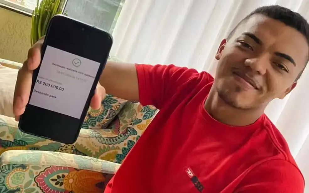 Homem em camiseta vermelha mostrando a tela do celular com uma notificação de Devolução realizada no valor de R$ 200 mil reais.