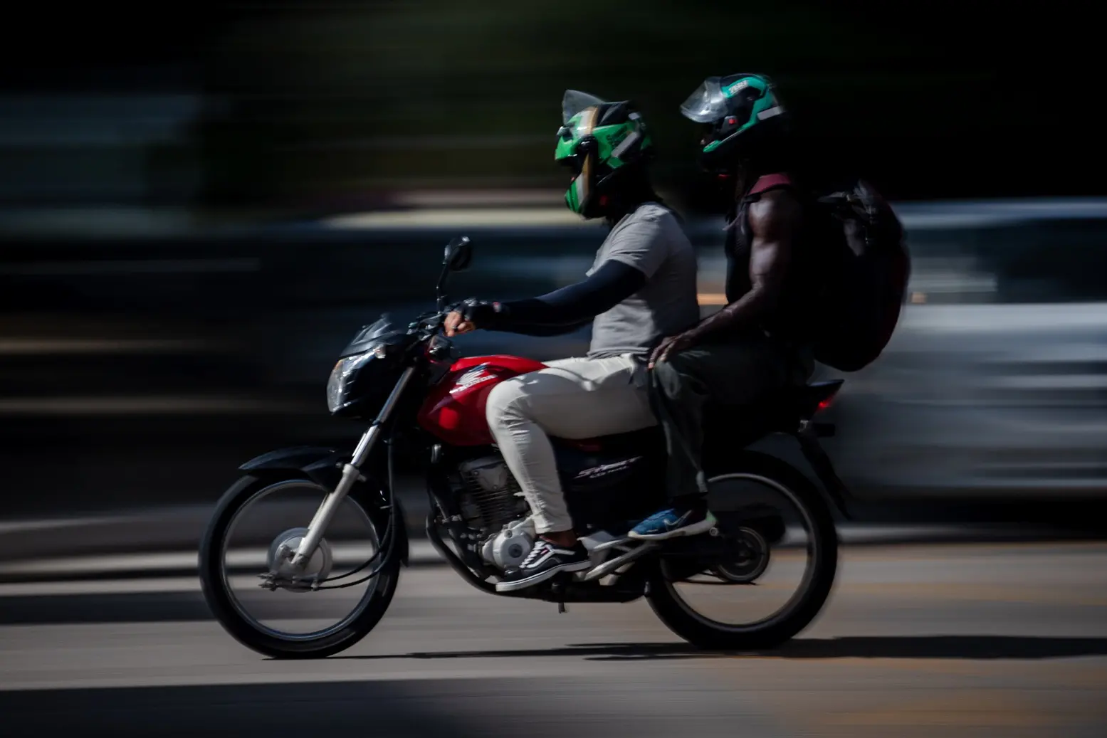 Imagem dinâmica de dois homens em uma moto vermelha, ambos usando capacetes verdes, com o cenário em borrão de movimento.