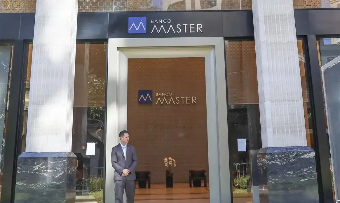 Fachada de uma agência do Banco Master, com duas grandes colunas claras nas laterais e um letreiro preto no topo exibindo o nome e o logotipo do banco. Na entrada, um segurança de terno cinza está parado em frente às portas de vidro. Dentro da agência, é possível ver uma parede com o logotipo do banco e alguns móveis decorativos.