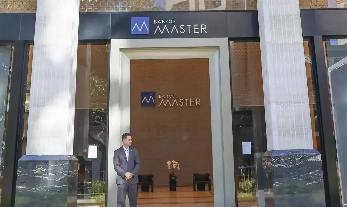 Fachada de uma agência do Banco Master, com duas grandes colunas claras nas laterais e um letreiro preto no topo exibindo o nome e o logotipo do banco. Na entrada, um segurança de terno cinza está parado em frente às portas de vidro. Imagem usada em matéria sobre Lula e Daniel Vorcaro.
