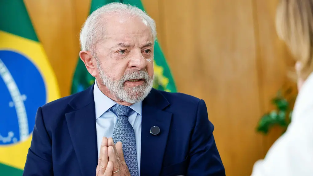 O presidente do Brasil, Luiz Inácio Lula da Silva, fala durante um evento oficial, vestindo terno azul com a bandeira brasileira no fundo.