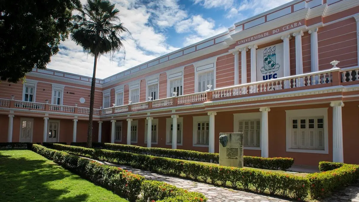 Edifício principal da Universidade Federal do Ceará (UFC) de cor rosa com colunas brancas, pátio ajardinado com palmeira e um busto memorial, sob céu azul claro.