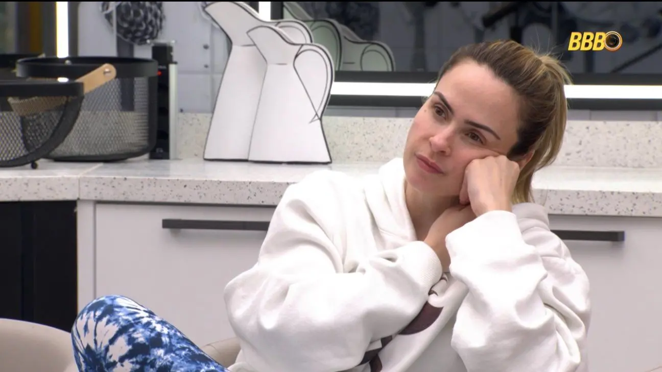 Ana Paula usando moletom branco com expressão pensativa, sentada em uma cozinha moderna com o logo BBB ao fundo.