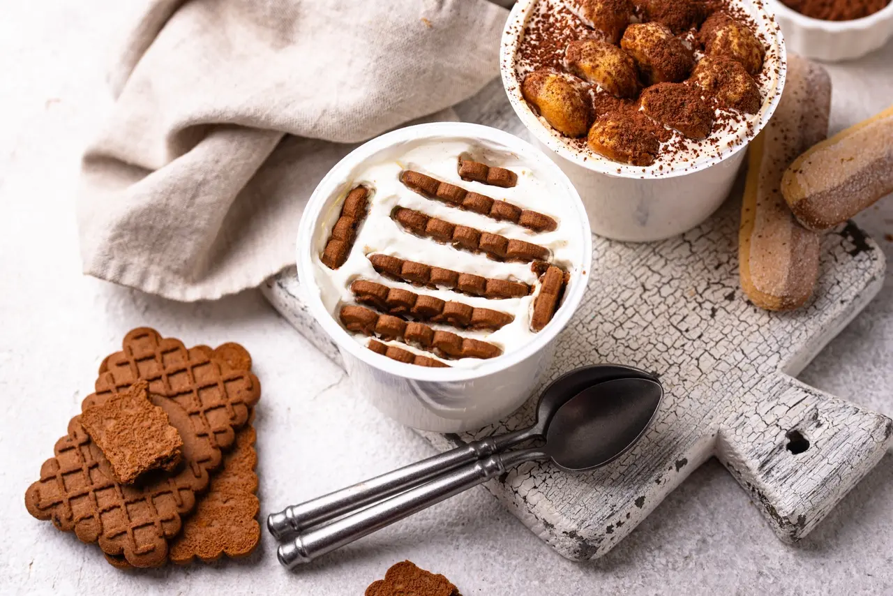Dois potes de sobremesa cremosa são apresentados sobre uma tábua rústica branca, com um deles destacando biscoitos de chocolate mergulhados em creme branco e o outro finalizado com cacau em pó. A composição vibrante inclui colheres de metal, biscoitos secos ao redor e um tecido de linho, criando um cenário gastronômico convidativo e detalhado.
