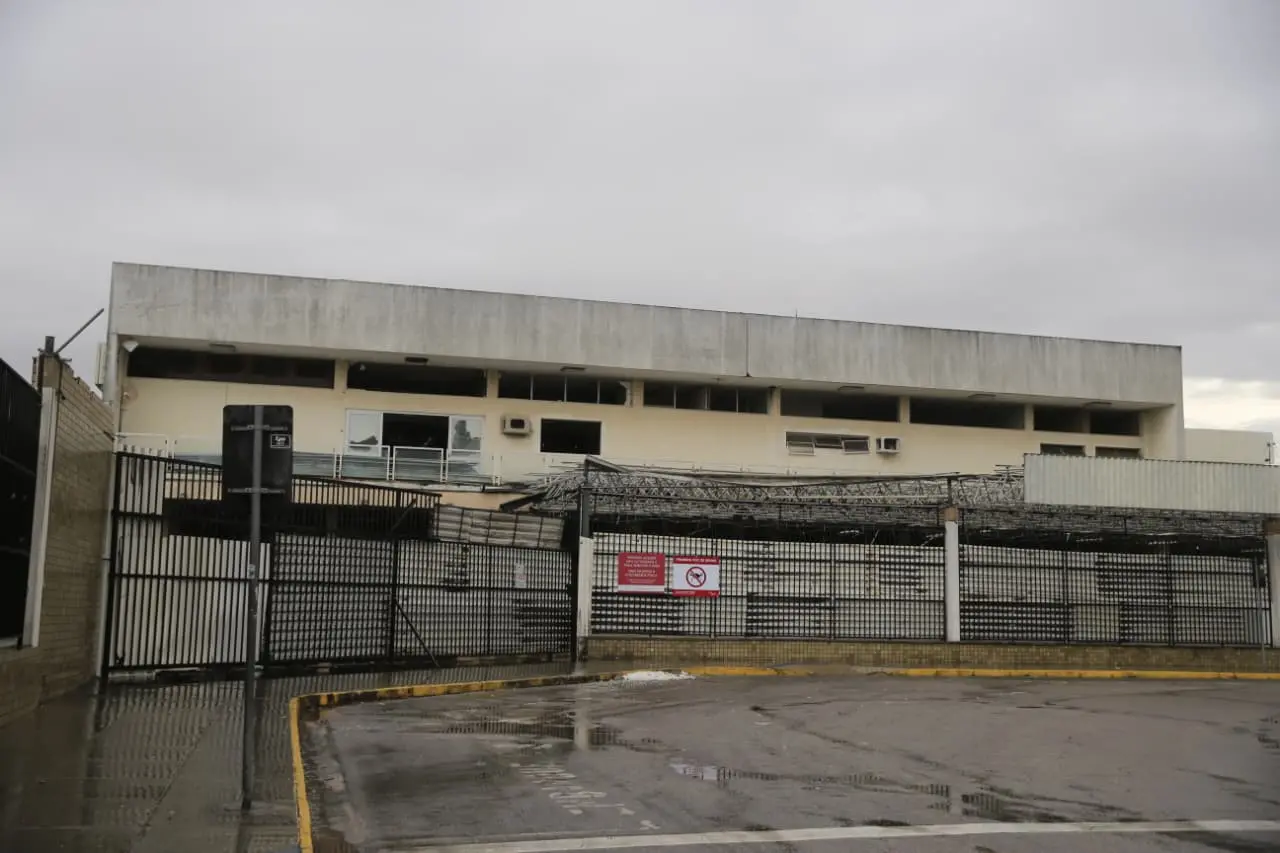 Antigo Aeroporto de Fortaleza. Estrutura retangular e o desgaste nas paredes externas. A área externa continua com marcas de chuva, e há poças d’água refletindo parte da fachada. O portão metálico ocupa boa parte da cena e exibe novamente a placa de proibição, sugerindo área restrita ou desativada.