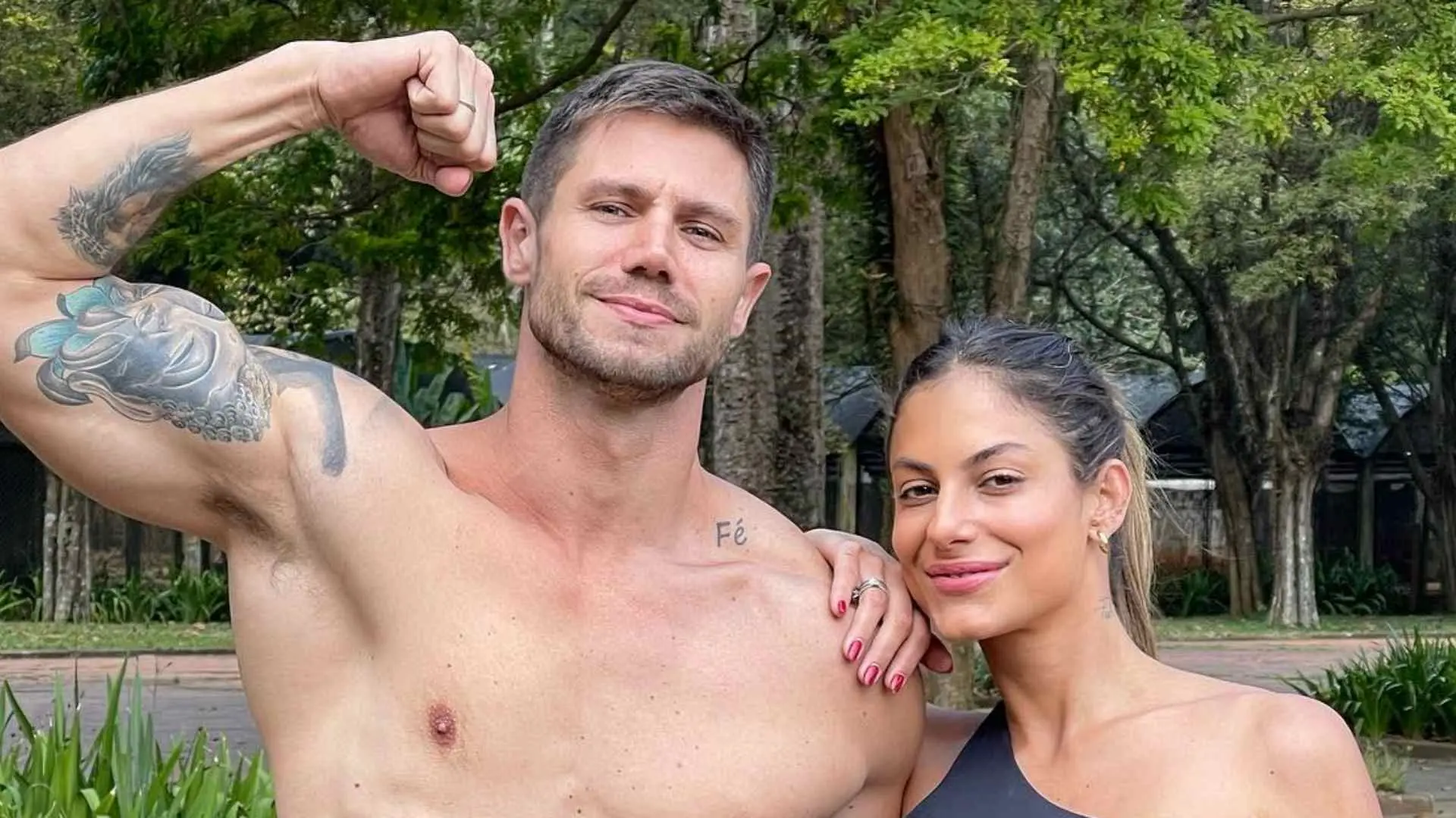 Um homem sem camisa posa ao ar livre, flexionando o braço esquerdo e exibindo uma tatuagem grande no bíceps. Ele está ao lado de uma mulher que veste roupa de ginástica escura e encosta a mão no ombro dele. Ambos estão em um ambiente arborizado, com árvores altas, vegetação e caminhos de um parque ao fundo.